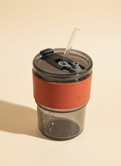 Insulated travel coffee mug with a reddish-orange leather grip and a clear reusable straw.