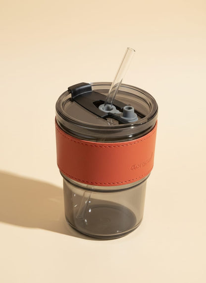 Insulated travel coffee mug with a reddish-orange leather grip and a clear reusable straw.